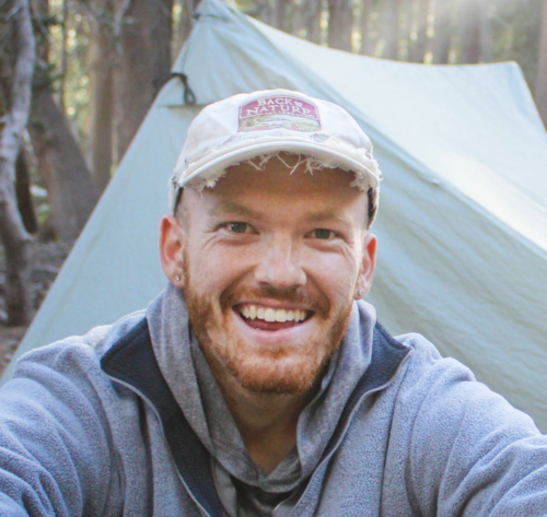 A portrait of me, smiling while sitting beside a trail in the woods.