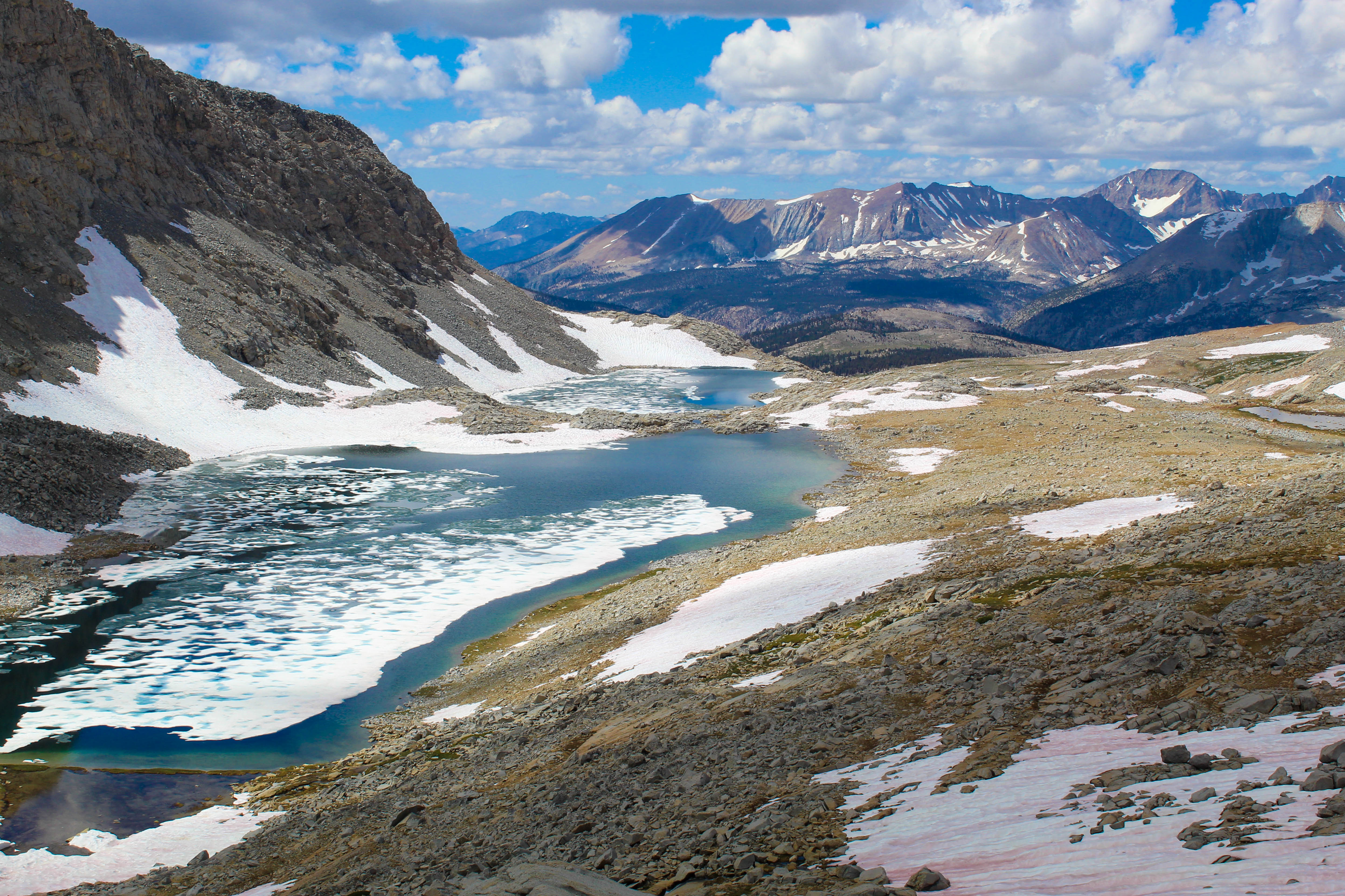 Two alpine lakes are half-covered in glaciers in the foreground. Behind, partly cloudy skies leave patterns of deep green and glowing green trees on the mountains in the distance.