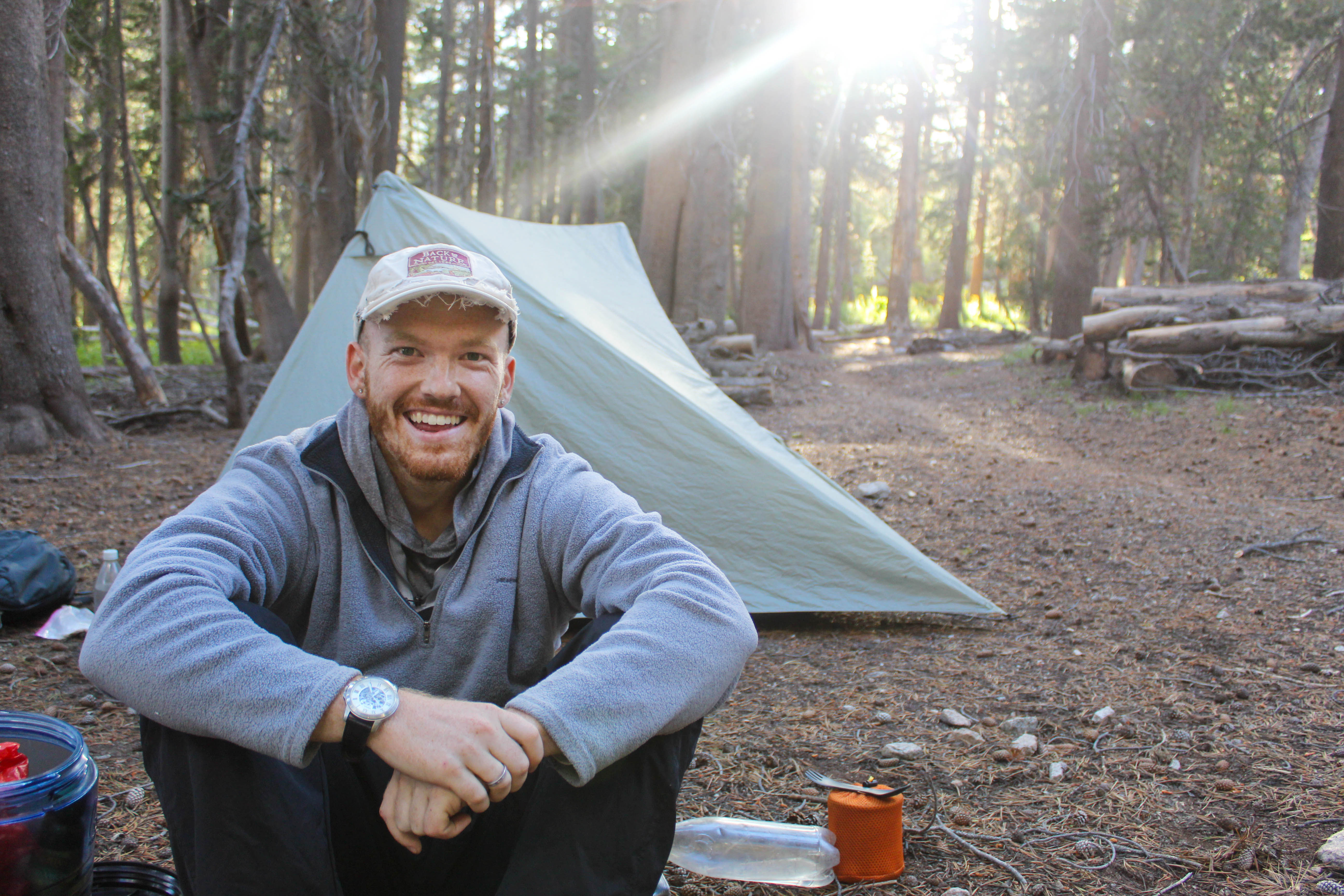 Me, sitting in front of a green tent beside the trail. Evening light shines through the trees behind.