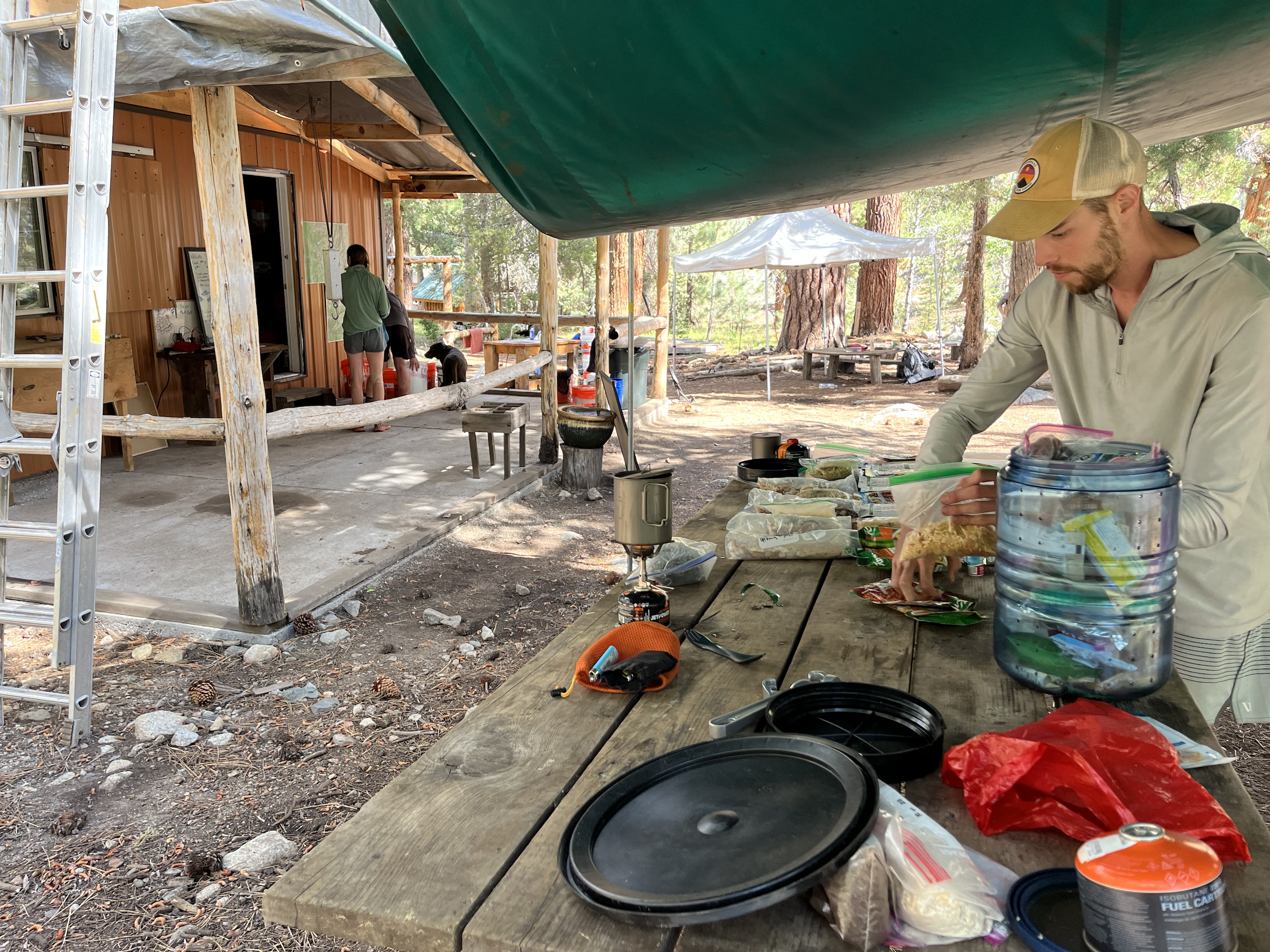 Chicken Legs packs his bear canister with his food scattered on a large wooden table. A small cabin in the cabin is littered with five-gallon buckets containing food and supplies.