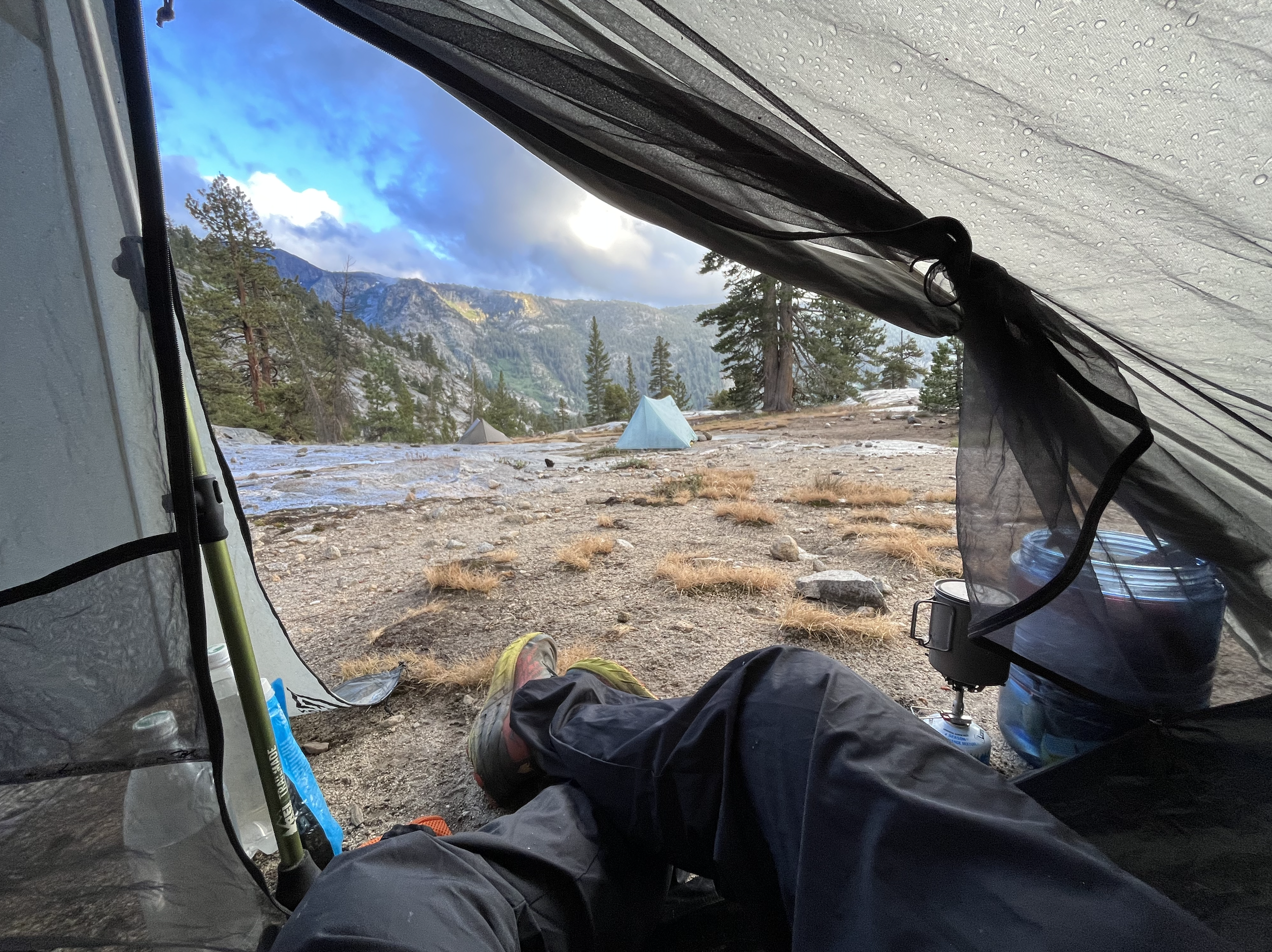 My legs reaching out into the post-storm evening light from inside the tent. Scattered tents and grey shrubs give way to jagged, partially lit peaks.